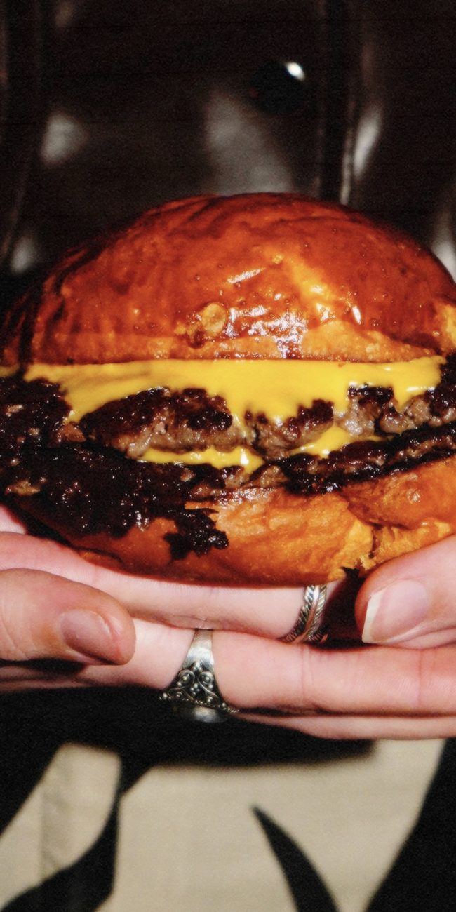 Junk: “Doble Smashed burger (240 gr.) de vaca rubia gallega. Madurada y salpimentada con un proceso de elaboración diario. Acompañada de queso cheddar y un crujiente bacon ahumado de la Sierra de Madrid. Chorreo de salsa JUNK. Envuelta en el brioche”.